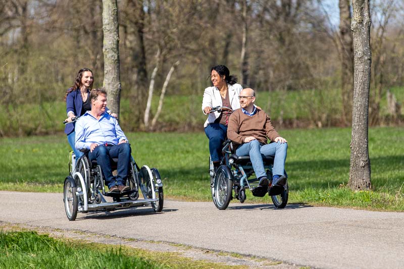 Zwei Männer werden in jeweils einem Rollstuhlfahrrad von zwei Frauen auf einem Feldweg befördert