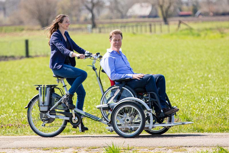Mann im Rollstuhl sitzt auf einem Rollstuhlfahrrad während eine Frau in die Pedale tritt